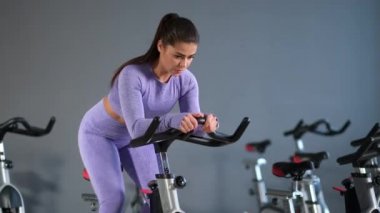 Spor kıyafetleri içinde güzel bir esmer kadın spor salonunda gri duvara karşı bisiklet sürüyor..