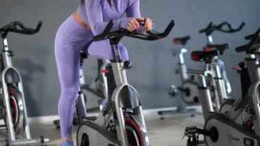 Spor kıyafetleri içinde güzel bir esmer kadın spor salonunda gri duvara karşı bisiklet sürüyor..