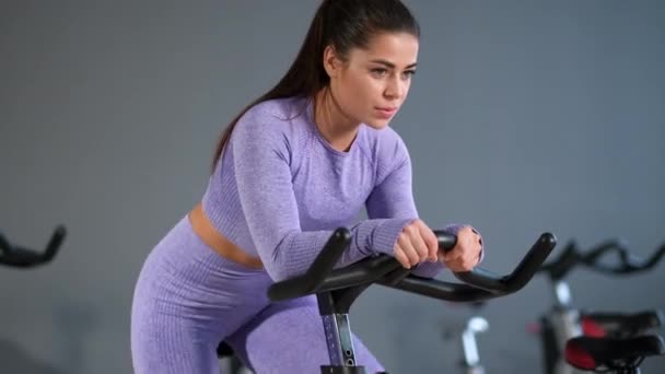 Une belle jeune femme brune sportive en vêtements de sport s'entraîne sur un sycle dans la salle de gym contre le mur gris. — Video