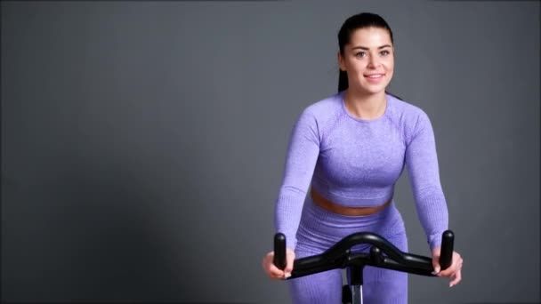 Una hermosa joven morena atlética en ropa deportiva entrena en un sycle en el gimnasio contra la pared gris. — Vídeos de Stock