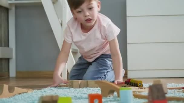 Um menino pré-escolar joga uma ferrovia de brinquedo de madeira no tapete da casa . — Vídeo de Stock