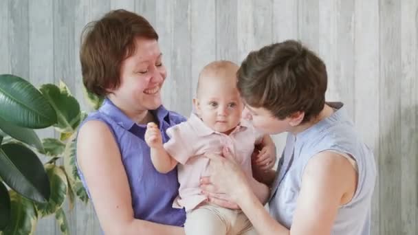 Två unga leende mödrar och en liten flicka. Mamma håller barnet i sina armar, kramar och kysser. Homosexuell lesbisk familj. — Stockvideo
