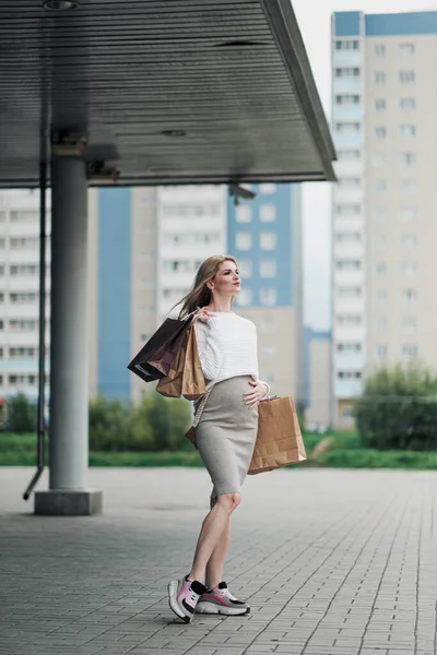 Jonge aantrekkelijke zwangere blonde vrouw in een witte trui tegen de achtergrond van de straten van de stad. Veel boodschappentassen vasthouden. — Stockfoto