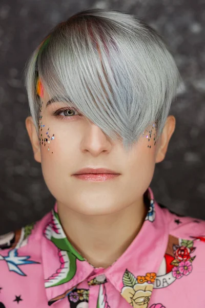 Portrait of a young beautiful girl in a pink shirt on a gray background with dyed hair. Short haircut pixie or bob. Rainbow coloring and sequins on the face. — Stock Photo, Image