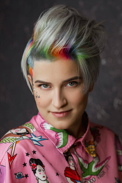 Portrait of a young beautiful girl in a pink shirt on a gray background with dyed hair. Short haircut pixie or bob. Rainbow coloring and sequins on the face.