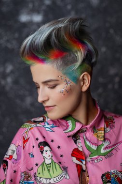 Pembe gömlekli, gri arka planda boyalı saçlı güzel bir kızın portresi. Kısa saçlı peri ya da kısa saçlı. Yüzünde gökkuşağı rengi ve pulları.