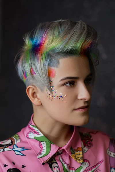 Portrait of a young beautiful girl in a pink shirt on a gray background with dyed hair. Short haircut pixie or bob. Rainbow coloring and sequins on the face. — Stock Photo, Image