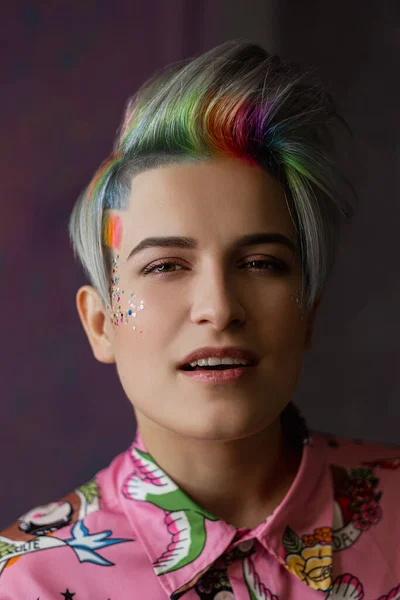 Portrait of a young beautiful girl in a pink shirt on a gray background with dyed hair. Short haircut pixie or bob. Rainbow coloring and sequins on the face. — Stock Photo, Image