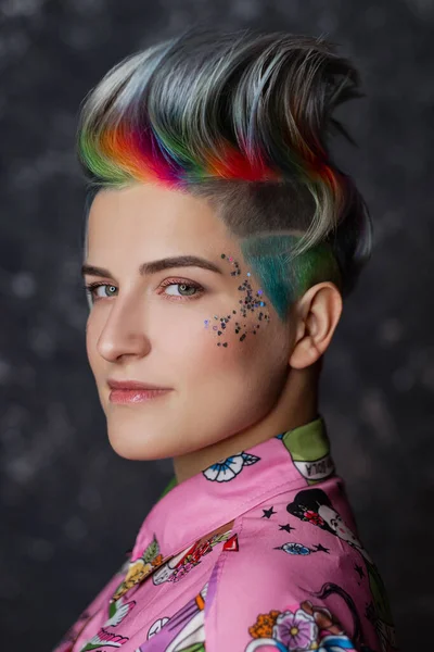 Portrait of a young beautiful girl in a pink shirt on a gray background with dyed hair. Short haircut pixie or bob. Rainbow coloring and sequins on the face. — Stock Photo, Image