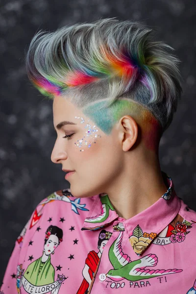 Retrato de uma jovem menina bonita em uma camisa rosa em um fundo cinza com cabelo tingido. Corte de cabelo curto pixie ou bob. Coloração do arco-íris e lantejoulas no rosto . — Fotografia de Stock