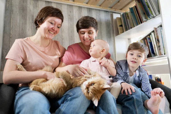 一个年轻的同性恋家庭，有两个孩子，一个儿子和一个女儿，他们花时间呆在家里。他们坐在沙发上看书。那只宠物猫躺在它旁边. — 图库照片