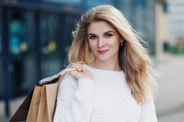 Beyaz kazaklı, çekici, hamile, sarışın bir kadın şehrin arka planında. Bir sürü alışveriş çantası taşıyorum..