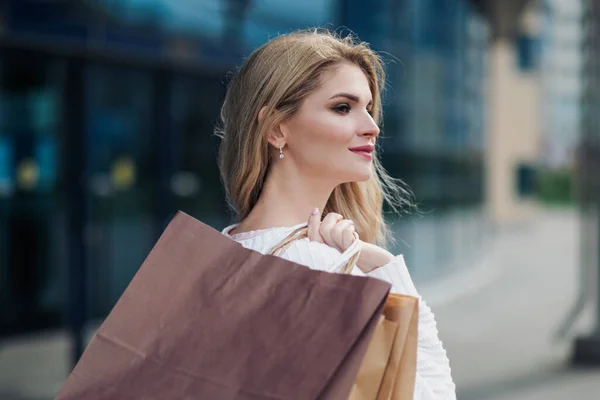 Jonge aantrekkelijke zwangere blonde vrouw in een witte trui tegen de achtergrond van de straten van de stad. Veel boodschappentassen vasthouden. — Stockfoto