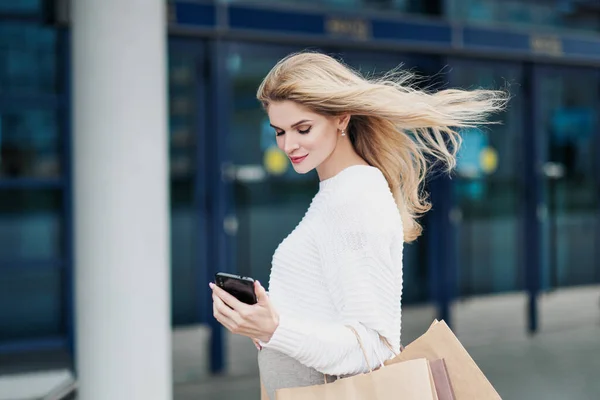 Jonge aantrekkelijke zwangere blonde vrouw in een witte trui tegen de achtergrond van de straten van de stad. Veel boodschappentassen vasthouden. — Stockfoto