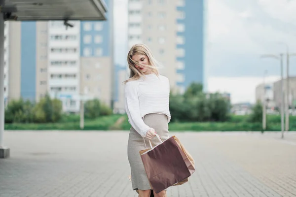 Jonge aantrekkelijke zwangere blonde vrouw in een witte trui tegen de achtergrond van de straten van de stad. Veel boodschappentassen vasthouden. — Stockfoto