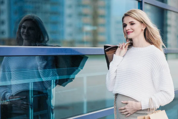 Joven mujer rubia embarazada atractiva en un suéter blanco en el contexto de las calles de la ciudad. Con un montón de bolsas de la compra . —  Fotos de Stock