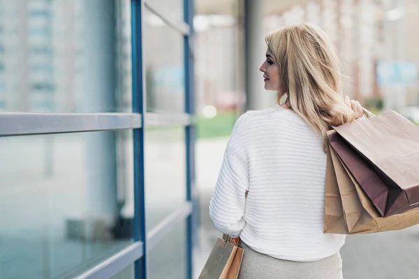 Jonge aantrekkelijke zwangere blonde vrouw in een witte trui tegen de achtergrond van de straten van de stad. Veel boodschappentassen vasthouden. — Stockfoto