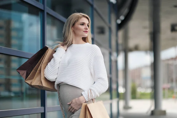 Jonge aantrekkelijke zwangere blonde vrouw in een witte trui tegen de achtergrond van de straten van de stad. Veel boodschappentassen vasthouden. — Stockfoto