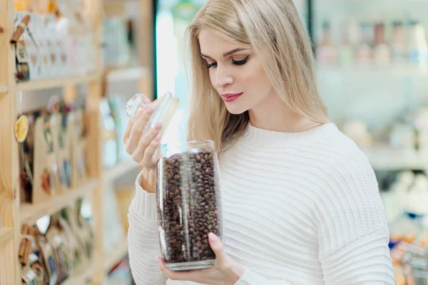 Une jeune femme blonde choisit le café aux céréales dans le magasin. Tests différentes variétés de café à partir d'un pot . — Photo