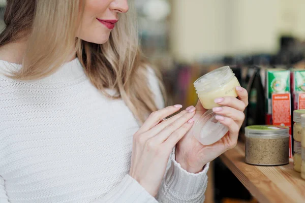 Une jeune femme blonde en pull blanc choisit la crème dans un magasin de cosmétiques . — Photo