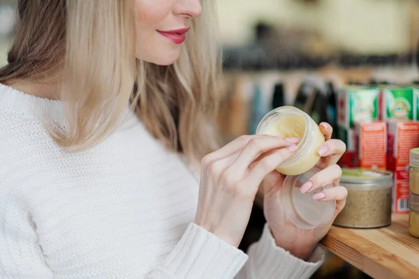 Une jeune femme blonde en pull blanc choisit la crème dans un magasin de cosmétiques . — Photo