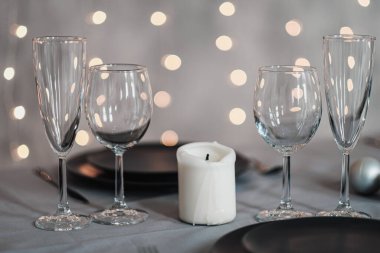 Restoranda bir masa ayarlandı. Masa servisi. Gri sanatsal arka plan. Siyah matte kaplar, cam bardaklar. Beyaz mumlar ve dekor. Arka planda şenlikli sarı ışıklar çelengi.