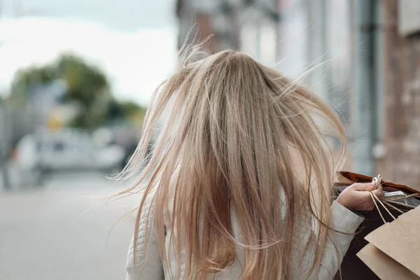 Joven hermosa mujer rubia está caminando a lo largo de una calle de la ciudad, el viento está soplando pelo largo. En las manos lleva una gran cantidad de paquetes con compras . —  Fotos de Stock