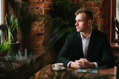 Yakışıklı, genç, sakallı, takım elbiseli ve gömlekli sarışın bir adam. Masaya oturur, dergi okur..