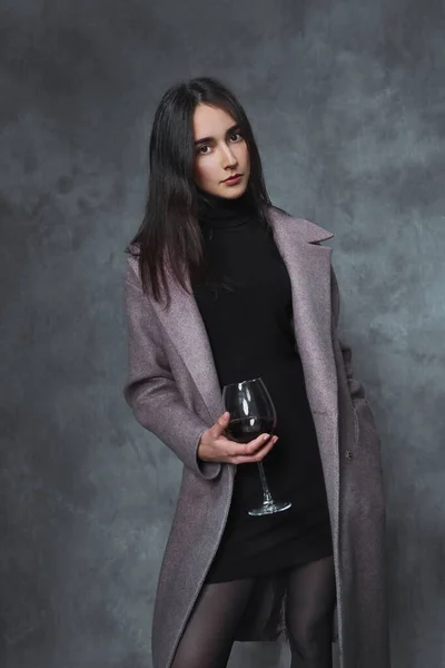 Красива молода брюнетка в чорному светрі і бузковому пальто на сірому фоні. келих червоного вина в руці . — стокове фото