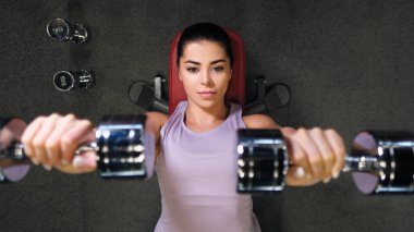 Spor kıyafetleri içinde spor salonunda güzel bir esmer kadın dambılları kaldırıyor..