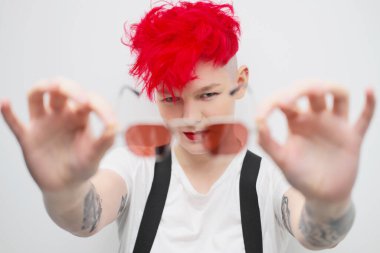 Havalı, modern genç bir kızın portresi. Tıraşlı şakağı olan kısa bir saç kesimi. Parlak kızıl saçlı. Kırmızı ruj. Beyaz arka planda stüdyo fotoğrafı. Pantolon askıları ve kareli pantolonlar.