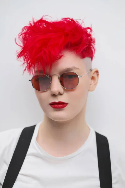 Retrato de uma jovem moderna e elegante. Um corte de cabelo curto com templo raspado. Cabelo vermelho brilhante tingido. Batom vermelho. Foto do estúdio sobre fundo branco. Suspensórios e calças quadriculadas . — Fotografia de Stock