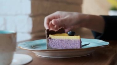 Siyah ceketli ve gözlüklü çekici genç bir kadın çatallı yaban mersinli cheesecake tatlısı yiyor..