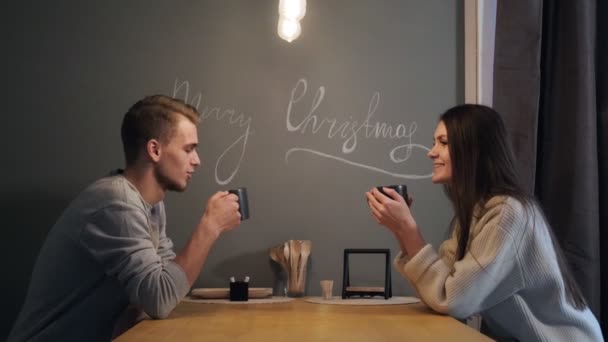 Jeune couple à la maison dans la cuisine du soir. Ils boivent du thé, rient et parlent. Une lampe jaune est allumée au-dessus de la table. Veille de Noël . — Video