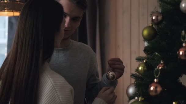 Ένα νεαρό ζευγάρι το βράδυ στολίζει ένα χριστουγεννιάτικο δέντρο στο σπίτι. Κρεμάστε μπάλες, αγκαλιά και γέλιο. Παραμονή Χριστουγέννων. — Αρχείο Βίντεο