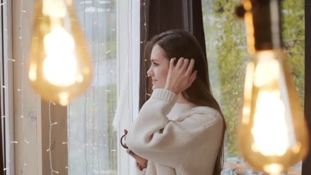 Portrait d'une jeune belle femme dans un pull chaud et avec une tasse de boisson chaude à la maison sur le fond d'une fenêtre. Lampes de lumière chaude . — Video