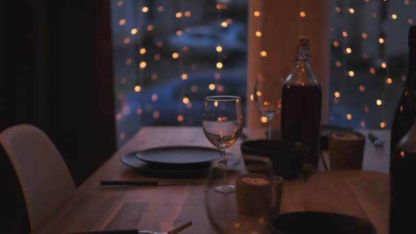 Mesa servida en casa en la sala de estar contra la ventana. Luz tenue de la noche. Las guirnaldas cuelgan de la ventana. Preparación para la celebración . — Vídeos de Stock