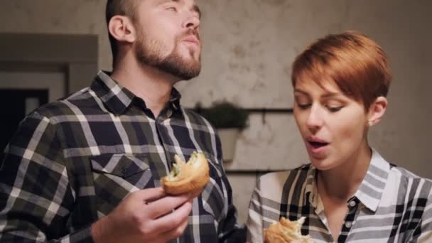 Joven pareja casada en ropa casual, hombre y mujer comiendo sándwiches en casa en la cocina. Luz nocturna de las lámparas amarillas . — Vídeos de Stock