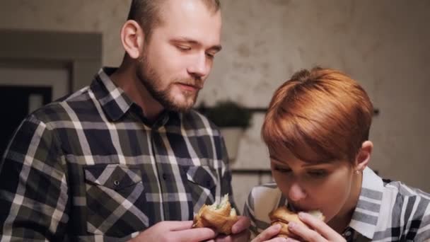 Giovane coppia sposata in abiti casual, uomo e donna mangiare panini a casa in cucina. Luce della sera dalle lampade gialle . — Video Stock