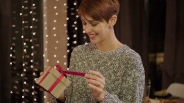 Evde hediye kutusuyla süveter giyen güzel, kısa saçlı bir kadın. Arka planda çelenk olan bir pencere var..