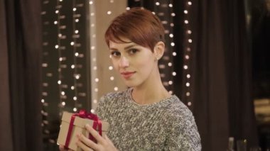 Evde hediye kutusuyla süveter giyen güzel, kısa saçlı bir kadın. Arka planda çelenk olan bir pencere var..