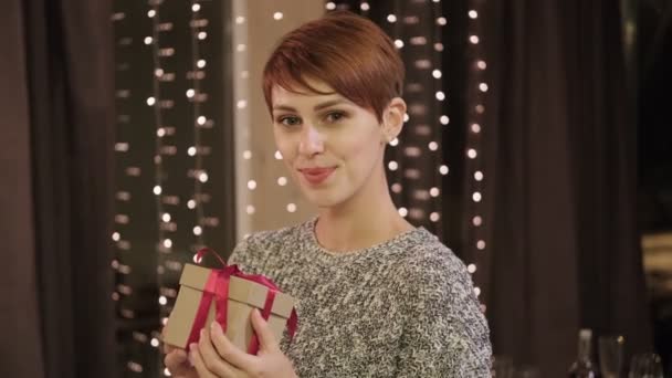 Junge schöne kurzhaarige Frau im Pullover mit Geschenkbox zu Hause vor dem Hintergrund eines Fensters mit Girlande. — Stockvideo