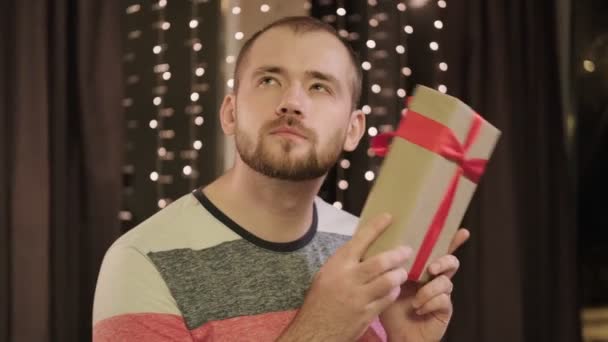 Young bearded man in casual clothes with a gift box at home against the background of a window with a garland. — Stock Video