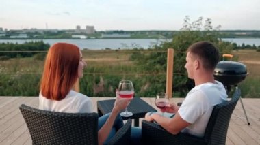 Genç, evli, beyaz bir çift terasta oturuyor ve şarap içiyor. Küçük, modern, ahşap bir evin arka planında. Barbekü ve şezlong sandalyeleri..