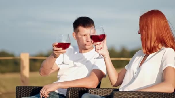 Una joven pareja caucásica casada se sienta en una terraza en sillones y bebe vino. Sobre el fondo de una pequeña casa de madera rústica moderna. Barbacoa y sillones . — Vídeos de Stock