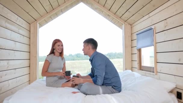 Jeune couple de famille caucasienne dans une petite maison rustique moderne avec une grande fenêtre. Allongé sur le lit, étreignant, jouant et regardant par la fenêtre . — Video