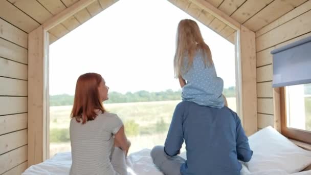 Junges kaukasisches Familienpaar mit kleiner Tochter in einem kleinen, modernen rustikalen Haus mit großem Fenster. Auf dem Bett liegen, sich umarmen, spielen und aus dem Fenster schauen. — Stockvideo