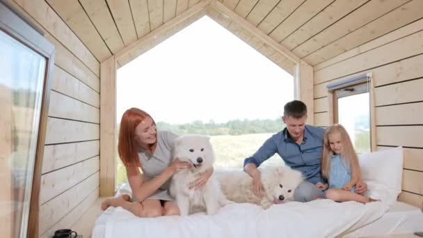 Jonge blanke familie echtpaar met dochtertje in een klein modern rustiek huis met een groot raam. Liggend op bed, knuffelend, spelend en kijkend uit het raam. Twee Samoyeds. Weekendje weg — Stockvideo