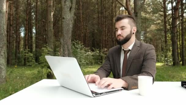 갈색 양복을 입고 넥타이를 매고 있는 매력적 인 젊은 남자 사업가는 책상에 앉아 바깥에 있는 컴퓨터로 일한다. 녹색 나무, 자연 및 공원의 배경. — 비디오