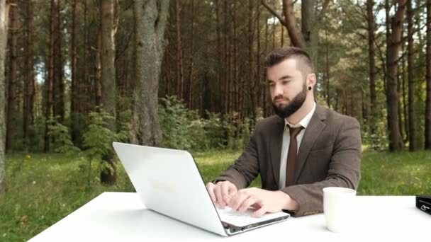 Joven atractivo hombre de negocios en traje marrón y corbata se sienta en el escritorio y trabaja en la computadora al aire libre. Árboles verdes, naturaleza y parque en el fondo . — Vídeo de stock